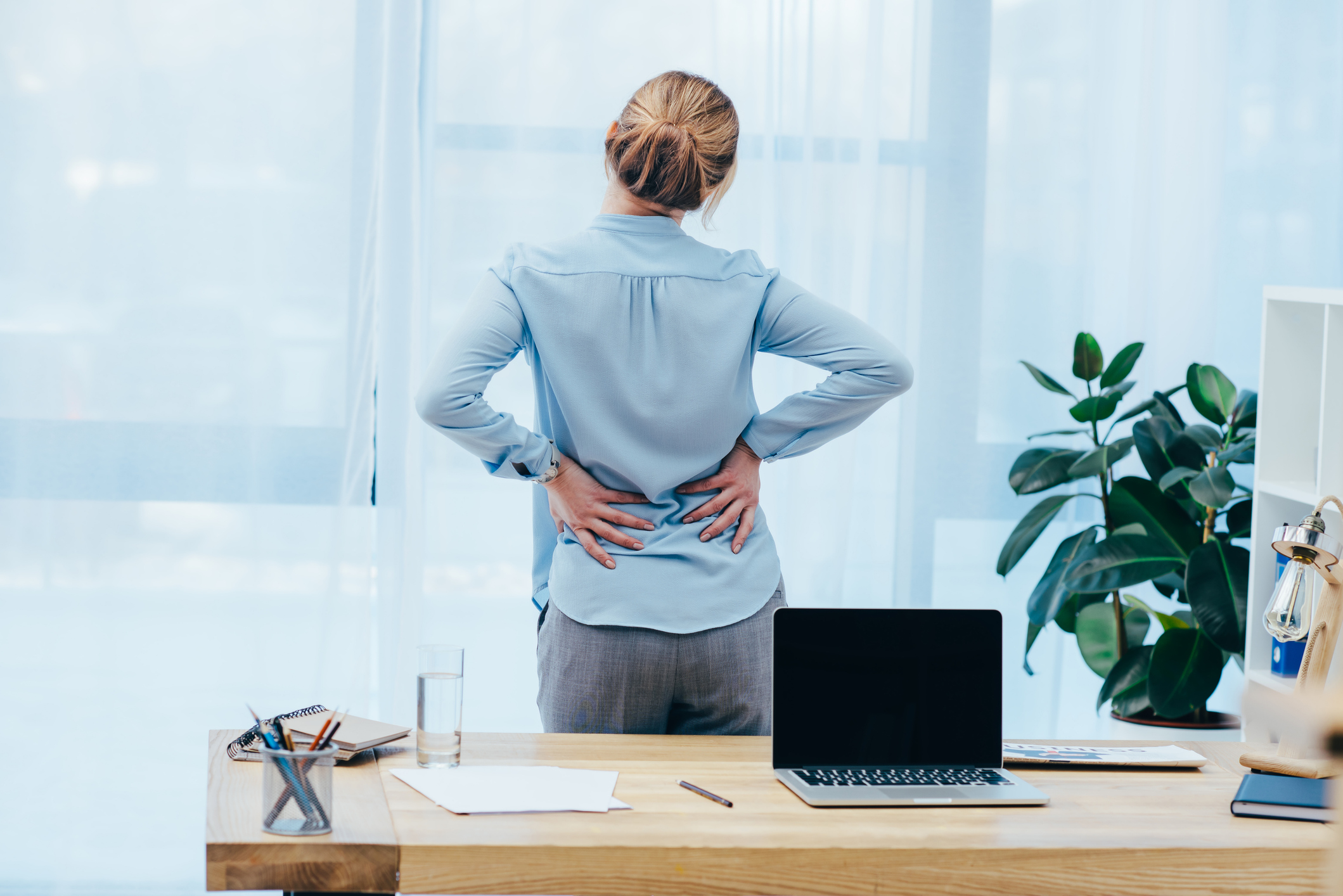 Woman experiencing back pain while standing at her desk, highlighting the need for services from a pain management clinic in Albuquerque.